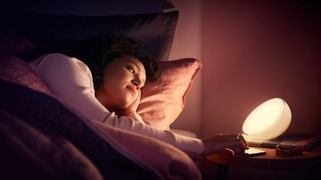 Een vrouw die in bed ligt, reikt naar het nachtkastje waar naast haar telefoon een tafellamp staat