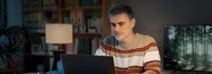 Un homme portant un pull rayé est assis à son bureau et regarde l'ordinateur portable devant lui.