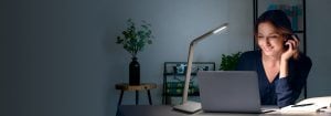 Une femme parle au téléphone tout en travaillant dans son bureau à domicile, avec une petite lampe de table à côté d'elle.