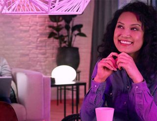 "Une lampe suspendue LED rose vibrante et une lampe à poser connectée créent une ambiance ludique, illuminant l'expression ravie de la femme dans un espace de vie moderne"